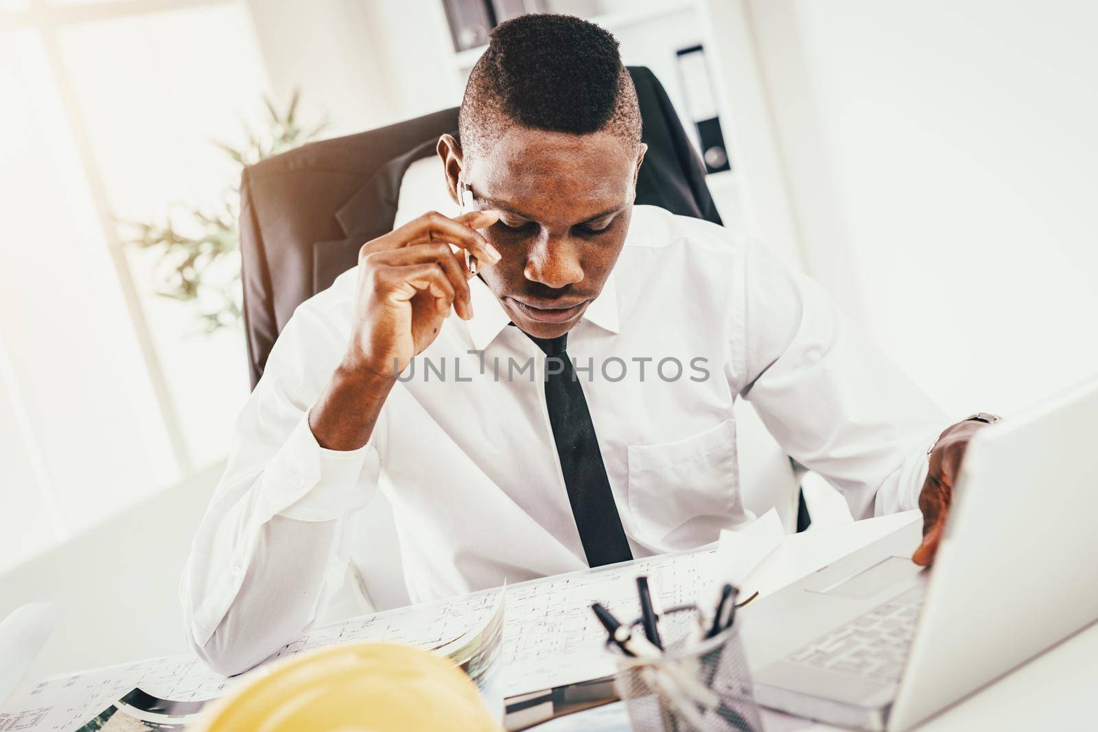 Pensive African architect analyzing blueprint and working on laptop in modern office and planning what to do next.