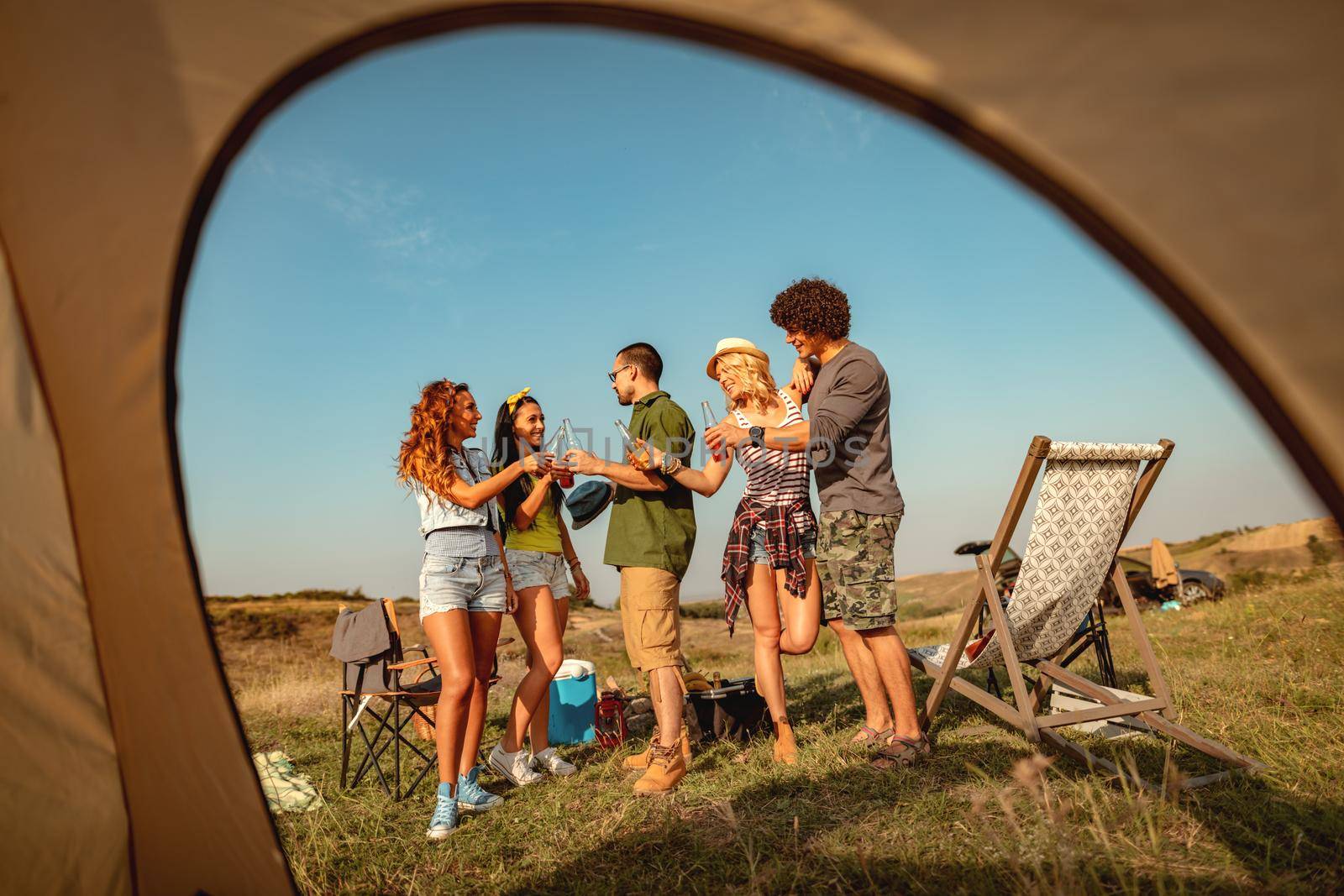 Happy young friends enjoy a sunny day in nature. They're clinking beer bottles, laughing and talking happy to be together.