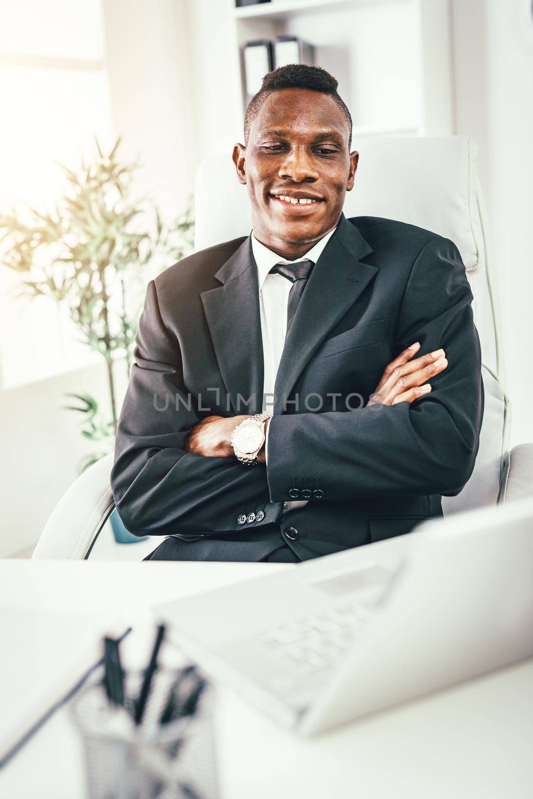 African smiling businessman sitting in the office with crossed arms and looking at laptop.