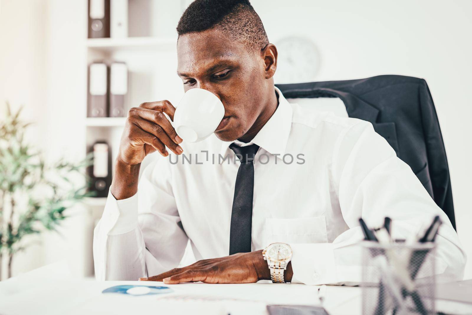 Pensive African businessman working in modern office, drinking coffe, reading  notes and planning what to do next.