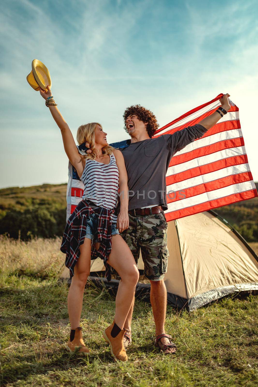 Happy young couple enjoys a sunny day in nature. They're hugging each other, holding an american flag in front a campsite tent.