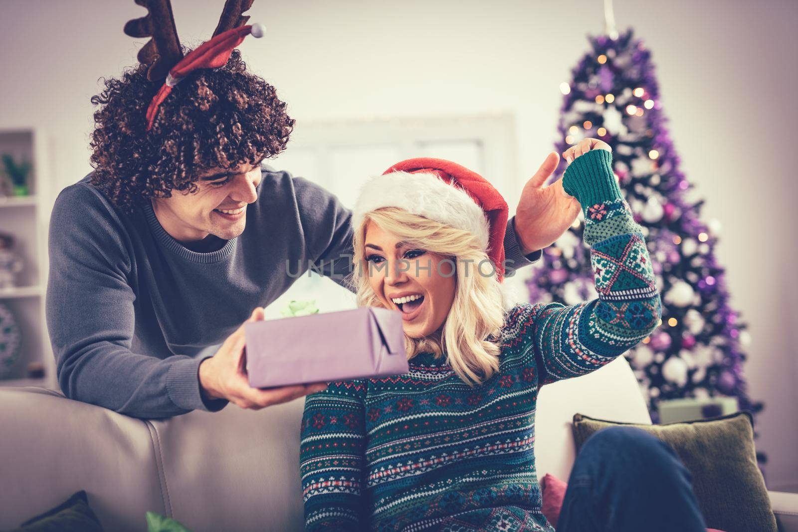 Beautiful young couple is celebrating Christmas at home. Handsome man is giving his girlfriend a gift box.