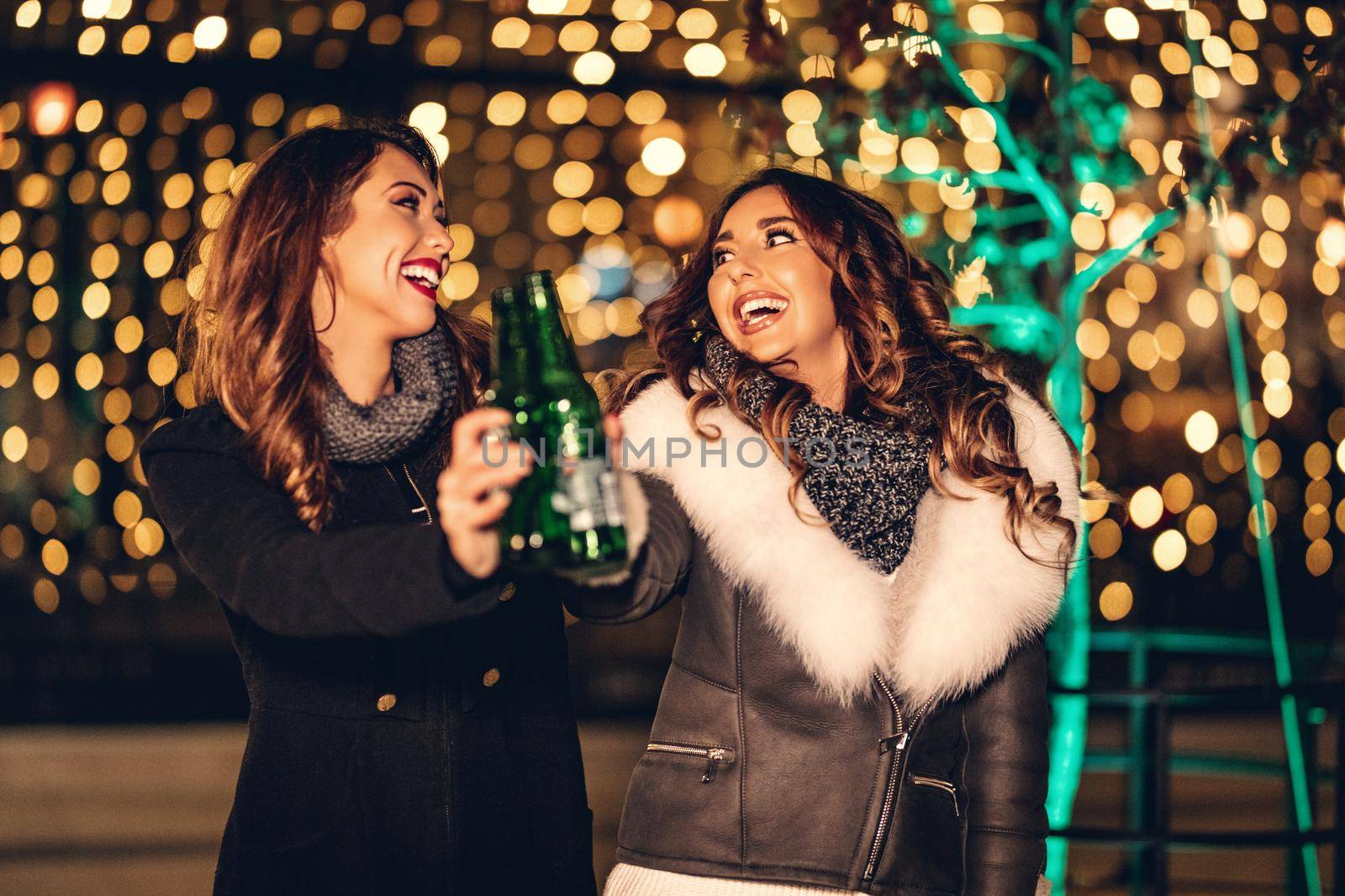 Two female friends enjoy the night out, looking each other and laughing with bottles of beer in their hands.