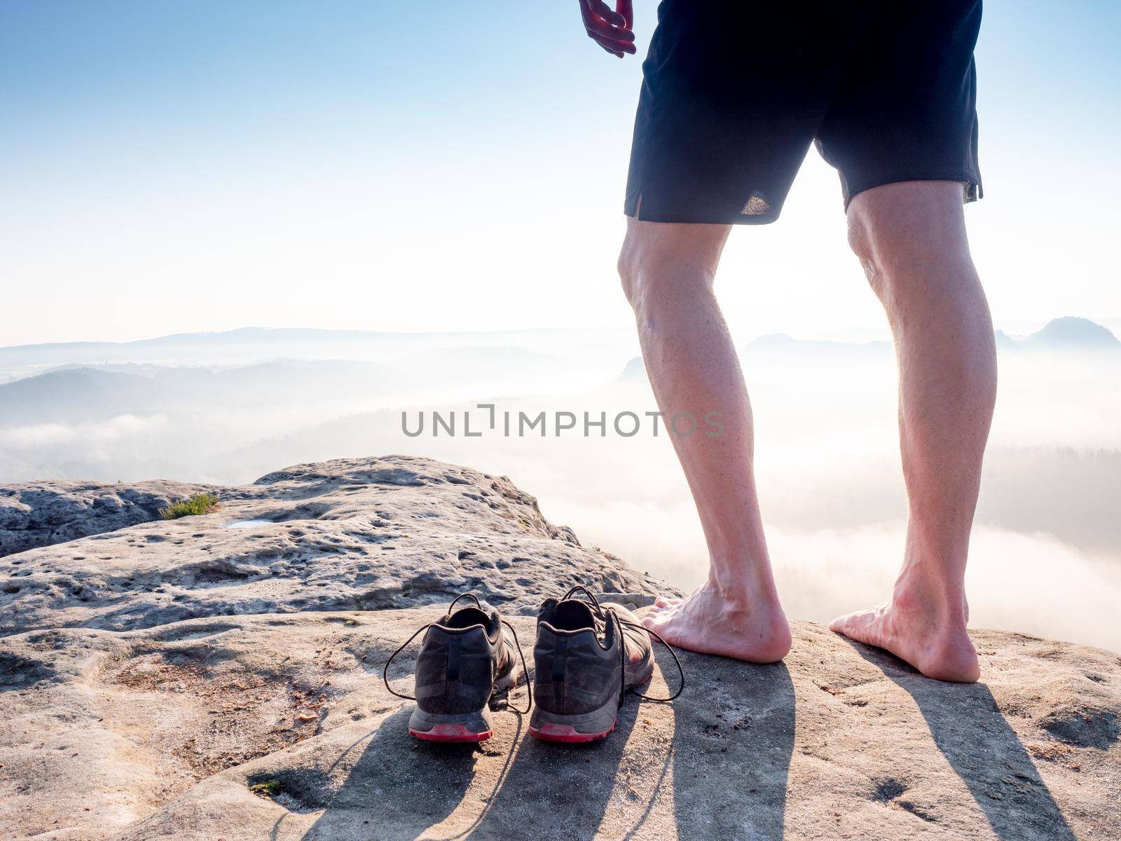 Traveler\'s legs, stripped light shoes and  mountain peak. Deep valley with creamy heavy fog and Sun hanging above horizon in distance