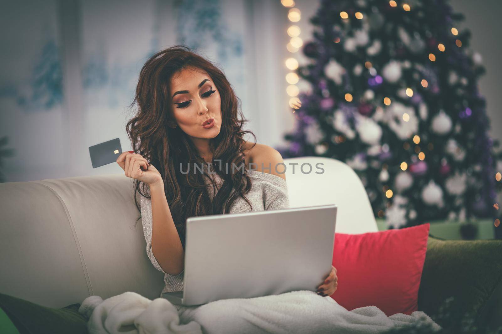 Cheerful young woman wrapped in a blanket and buying Christmas presents online at home.