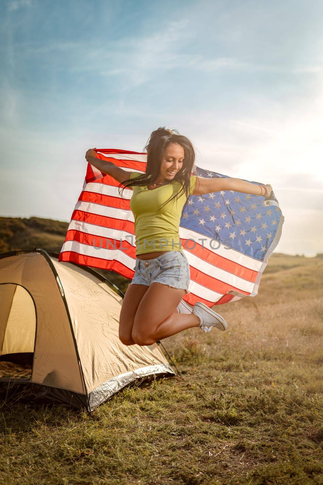 Happy young woman enjoys a sunny day in nature. She's holding an american flag in front a campsite tent and jumping of happyness. 
