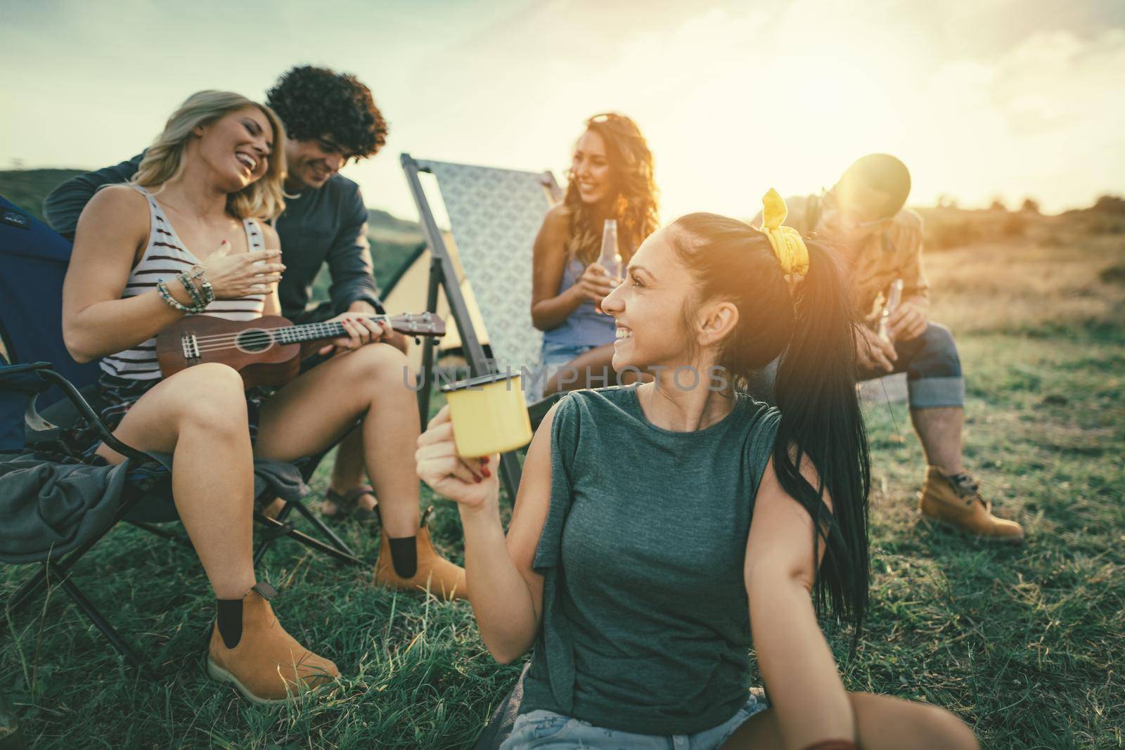 Happy young friends enjoy a sunny day at the mountain. They're laughing and singing with music from ukulele near tent.