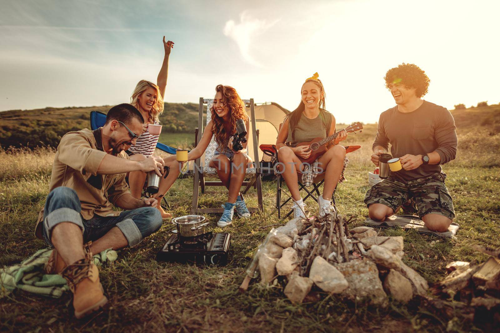 Young people have a good time in camp in nature. They're resting, drinking tea, laughing and singing with music from ukulele, happy to be together.