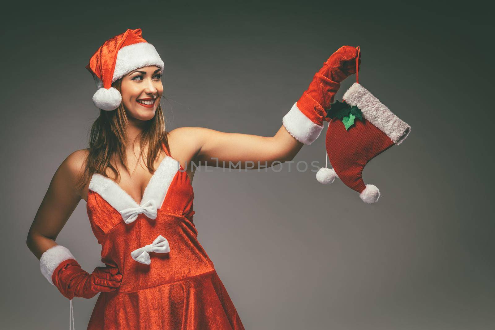 Beautiful young smiling woman in Santa Claus costume thinking and holding red christmas stocking.