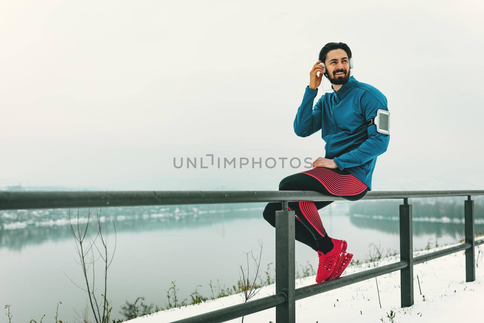 A male runner with headphones on his ears taking a break in the public place during the winter training outside beside the river. Copy space.