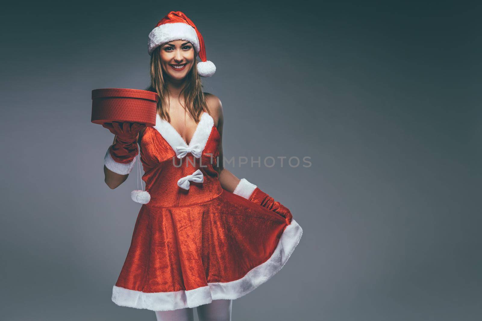 Beautiful young smiling woman in Santa Claus costume holding red present and looking at camera.