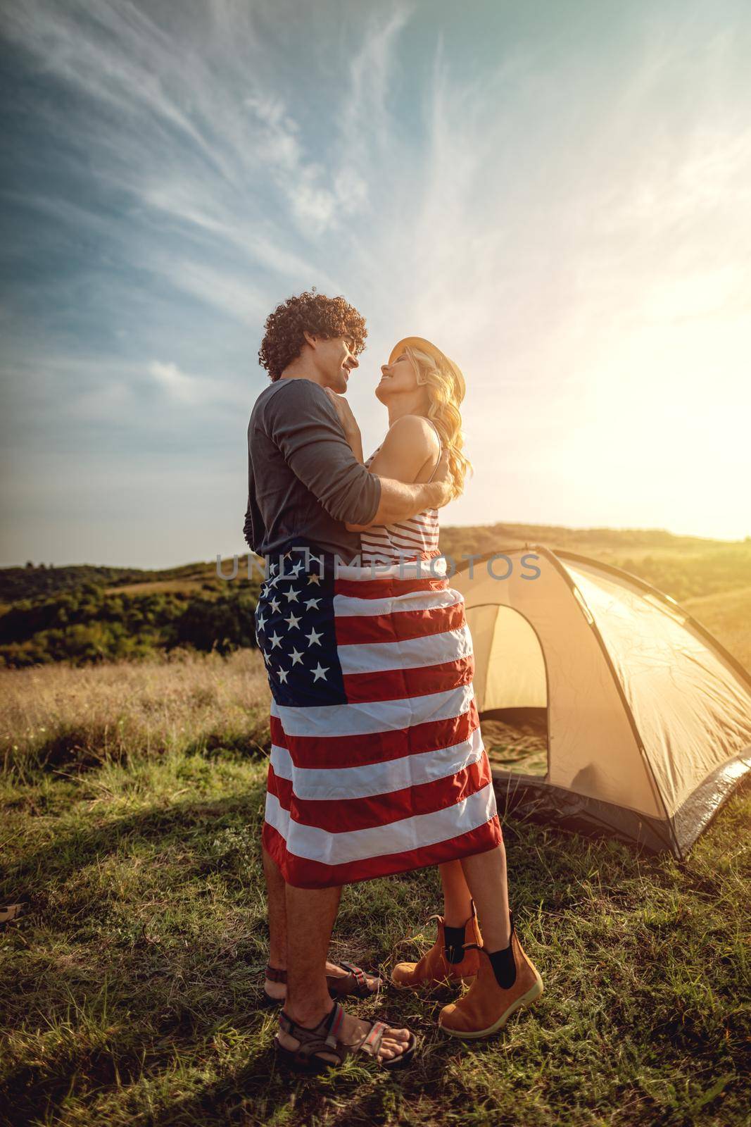Happy young couple enjoys a sunny day in nature. They're hugging each other, wrapped in american flag in front a campsite tent. 