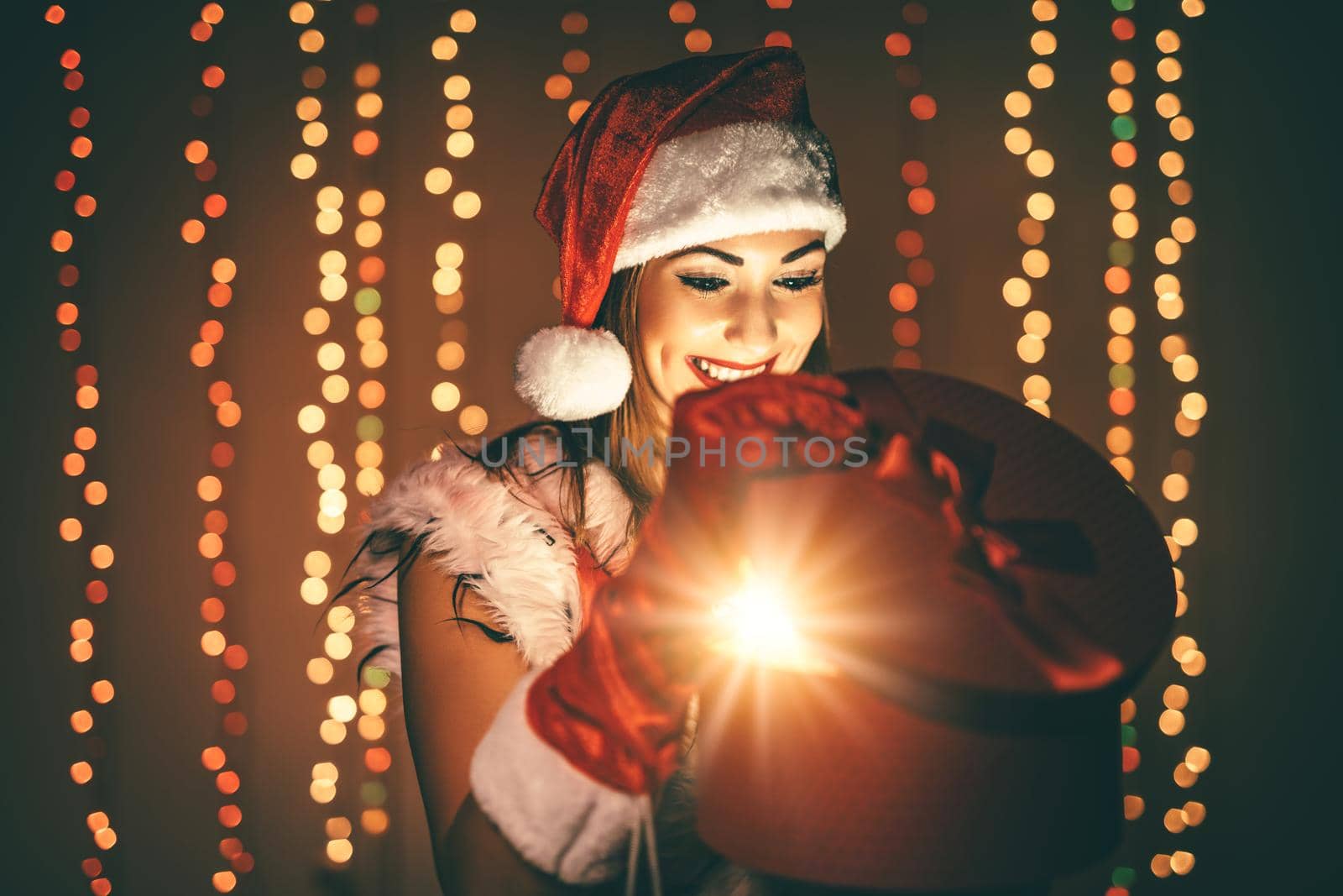Beautiful young smiling woman in Santa Claus costume holding red present and having fun looking what is in it..