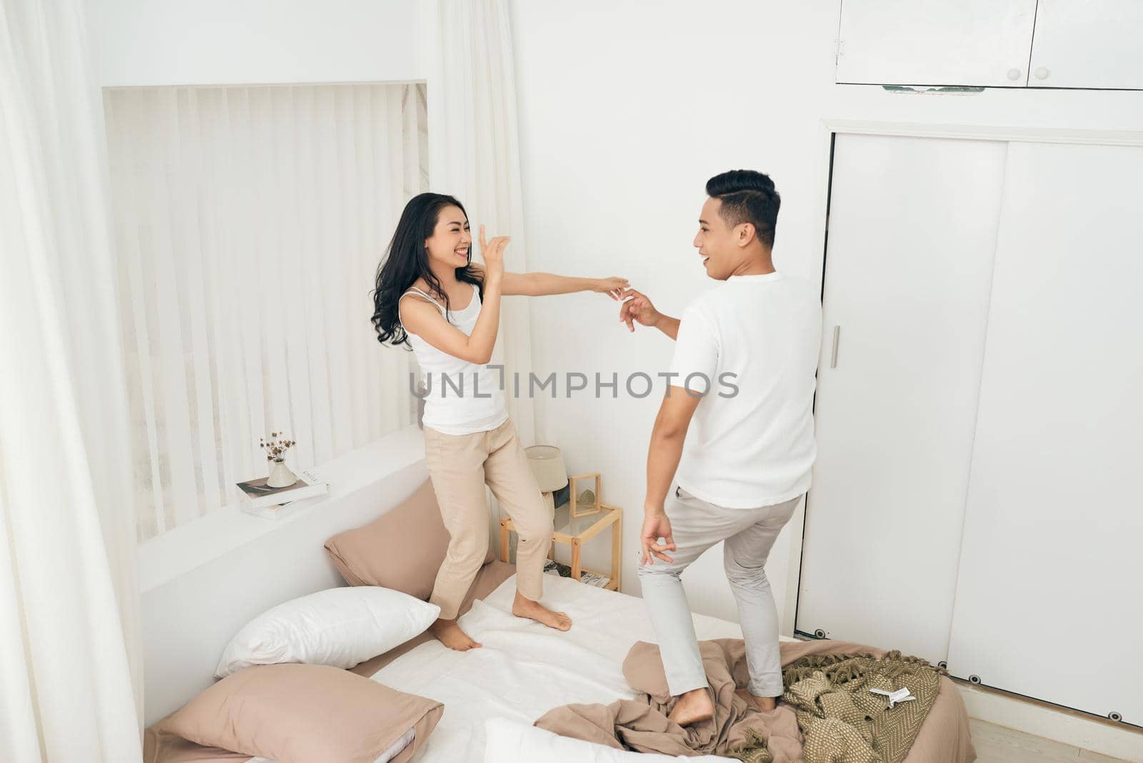 Dancing on the bed. Full length of beautiful young couple holding hands and smiling while dancing on the bed at home by makidotvn