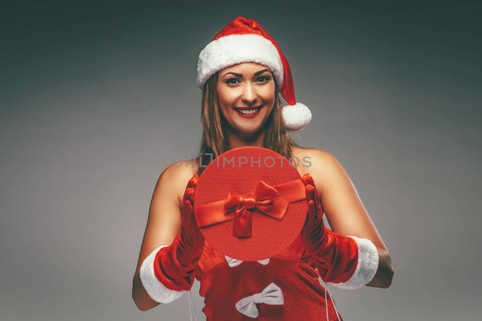 Beautiful young smiling woman in Santa Claus costume holding red present and looking at camera.