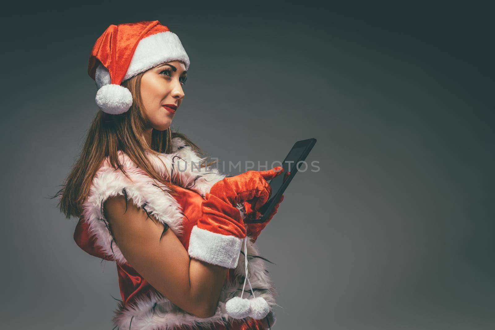 Pensive cute girl in Santa Claus costume making a list presents on a digital tablet.