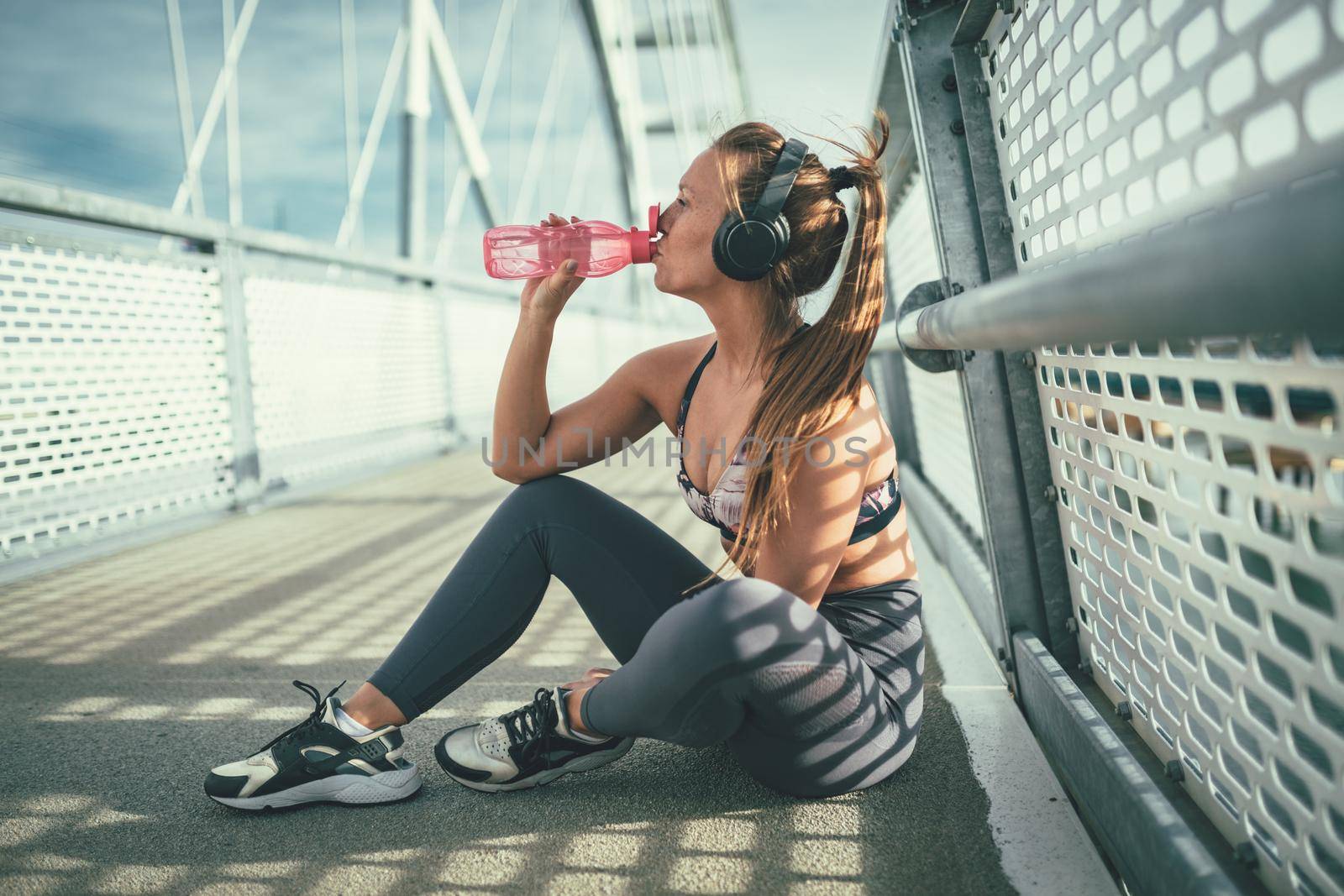 Young fitness woman is resting after hard training on the river bridge and drinking water.