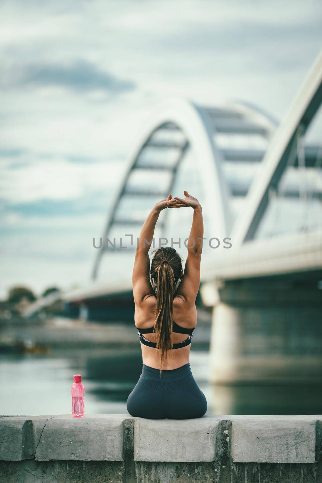Young fitness woman is resting and stretching after hard training by the river bridge, drinking water.