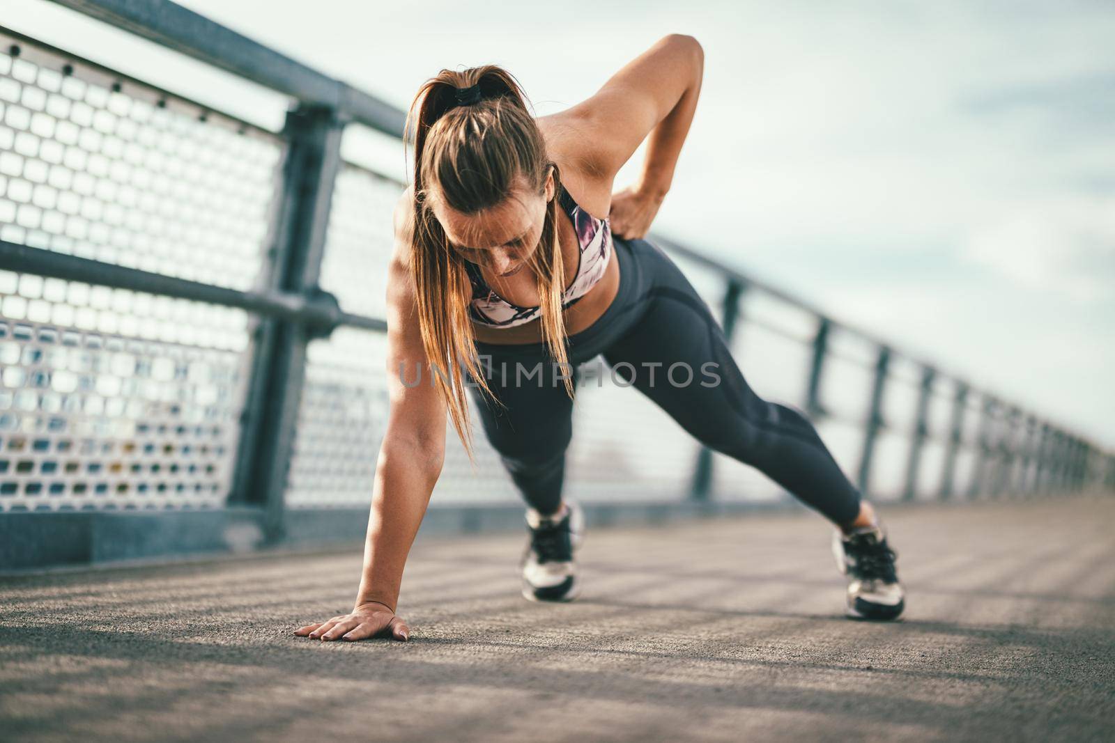 Young female runner doing push-upos exercise on a river bridge, preparing for morning workout.