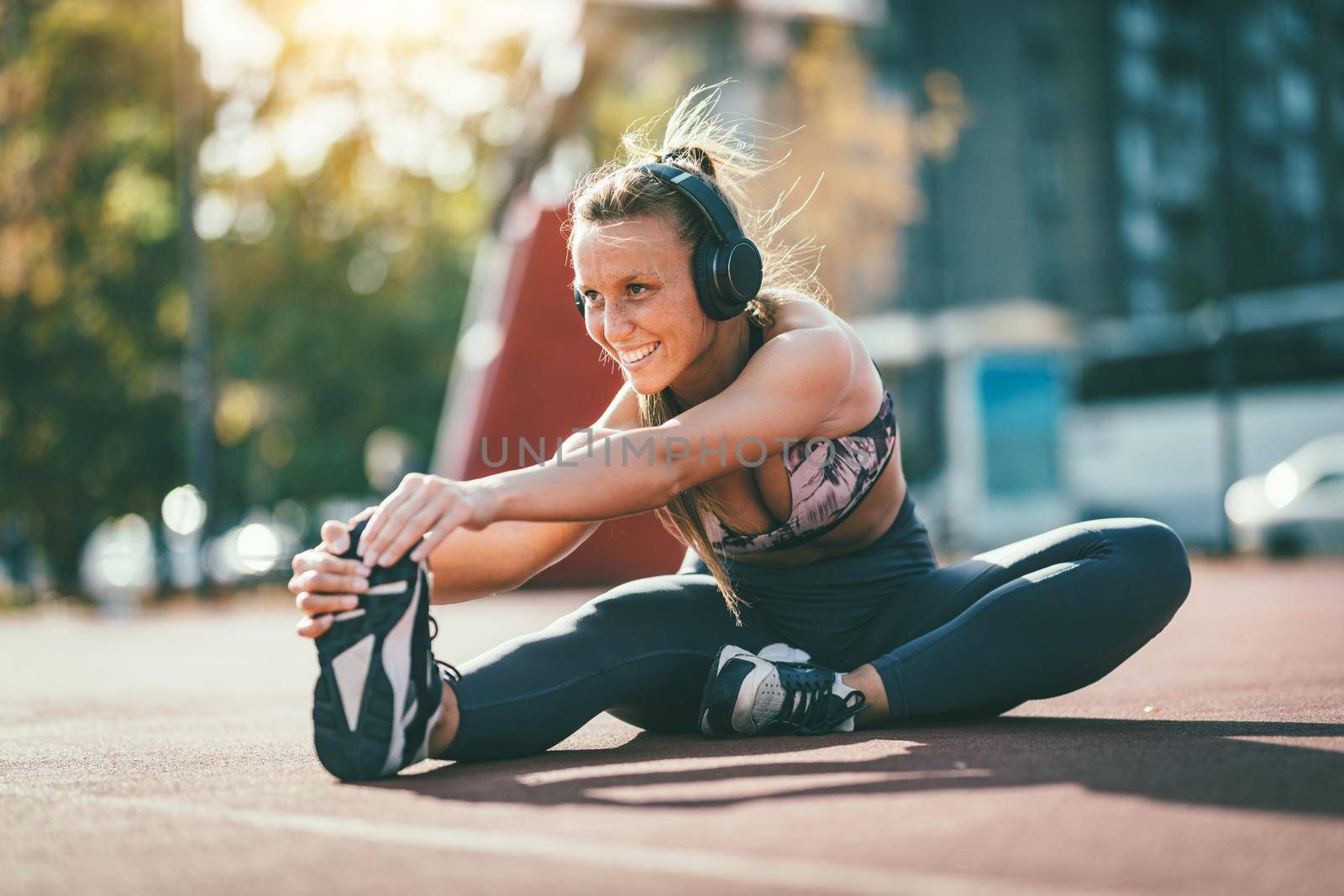 Young female runner, with headphones on her ears, doing stretching exercise on a public place, preparing for morning workout.