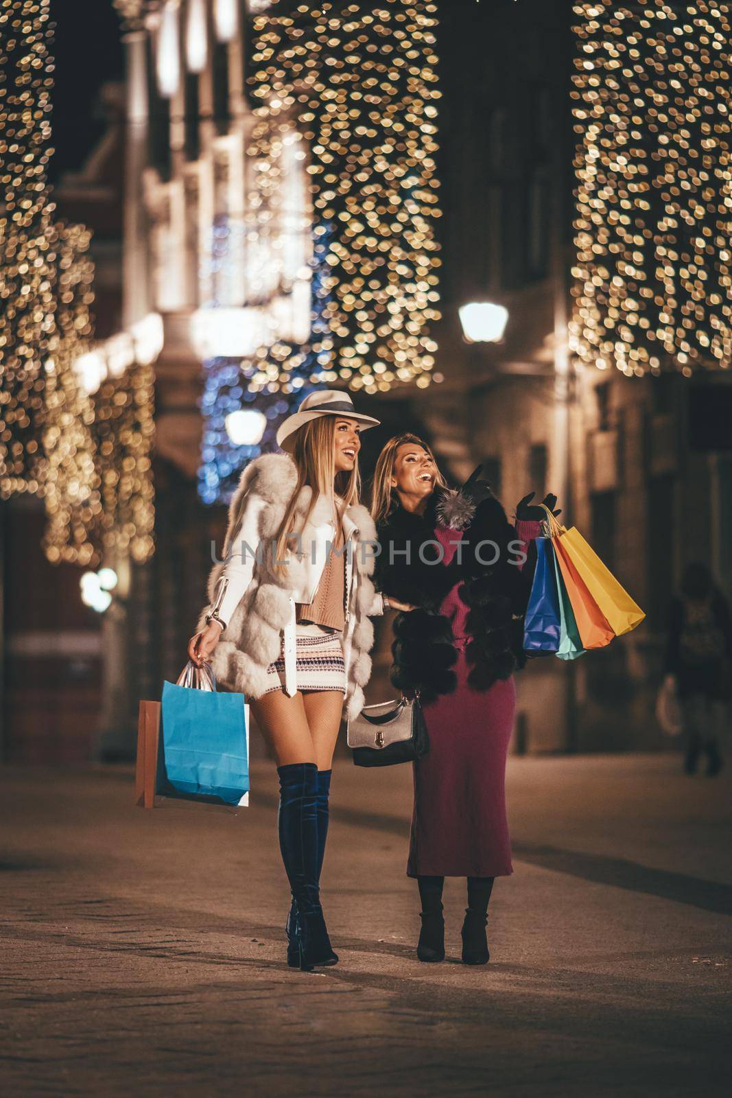 Young beautiful two cheerful sisters, with colorful shopping bags, having fun and walking in the night city street at Christmas time.