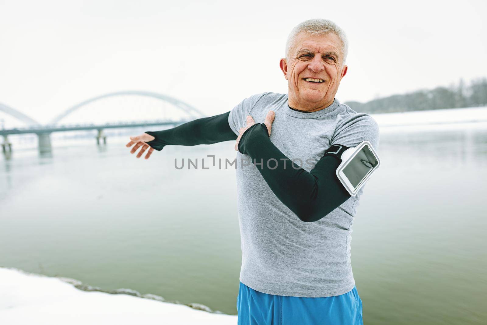 Active senior man stretching and doing exercises by the river during the winter training outside in.