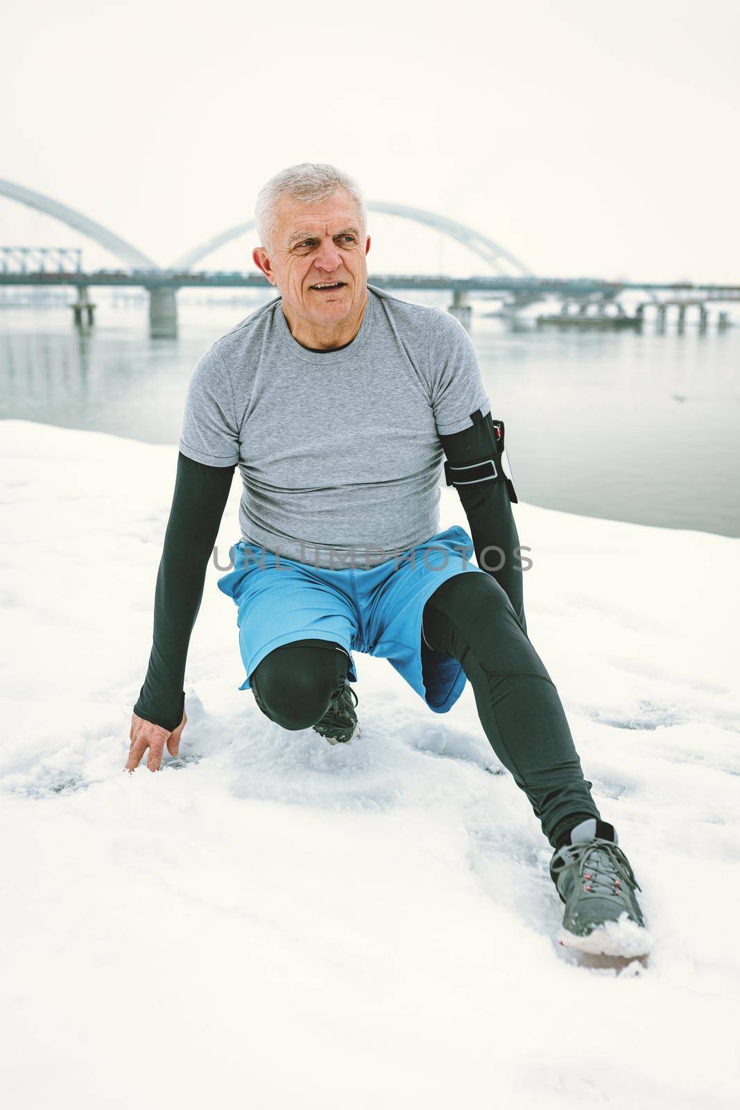 Active senior man stretching and doing exercises by the river during the winter training outside in.