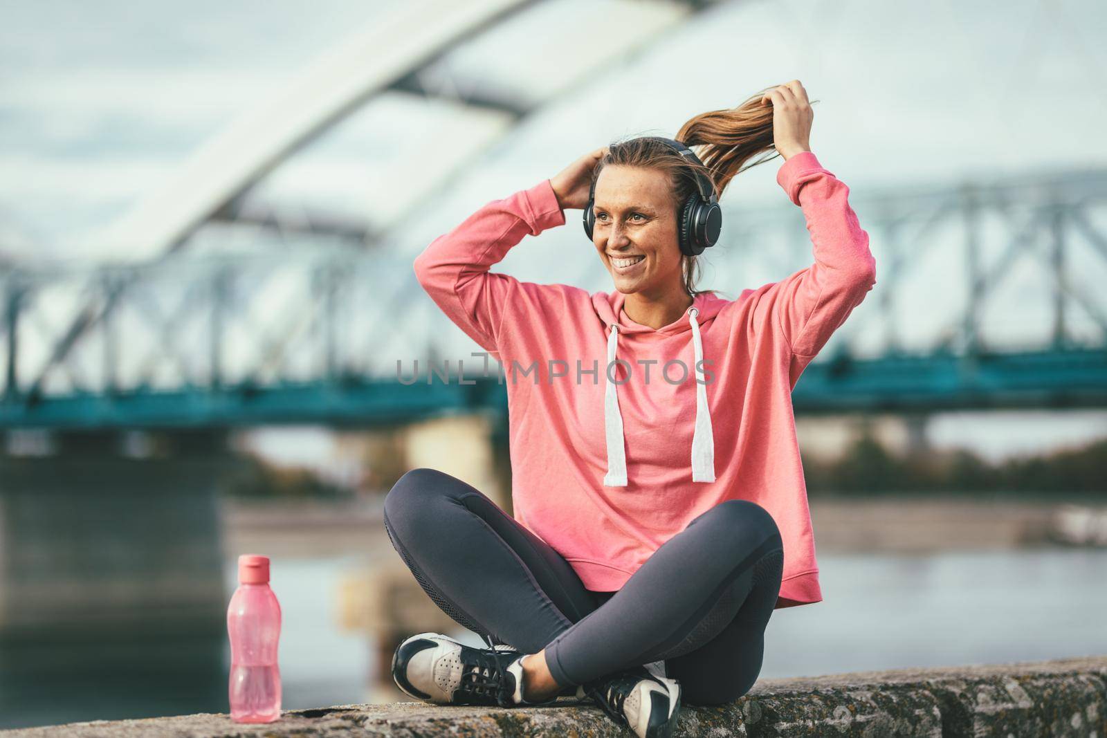 Young fitness woman with headphones is resting after hard training on the wall by the river with bottle of a water.