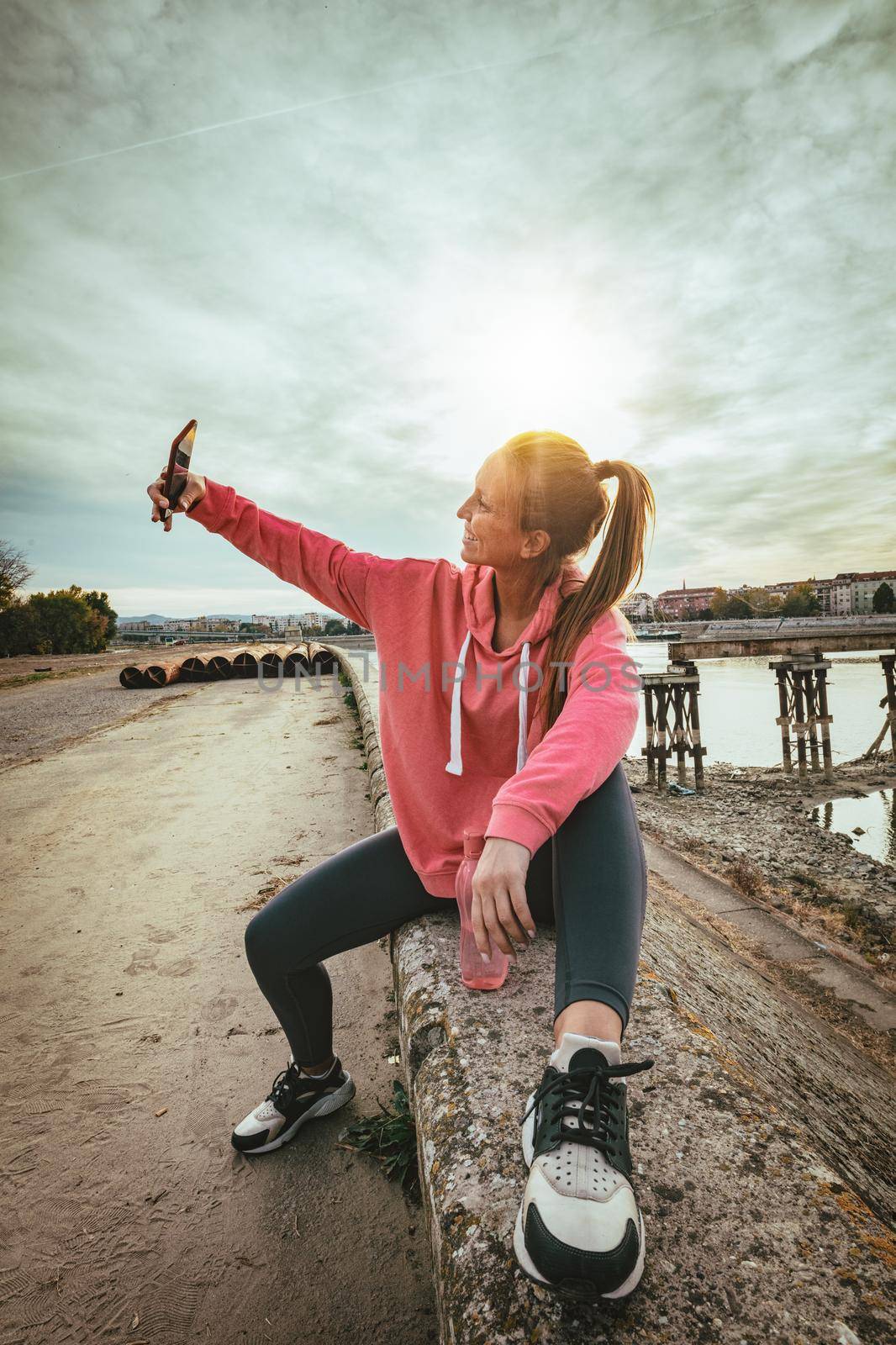 Young fitness woman is resting after hard training on the wall by the river bridge and taking selfie with smarthphone.