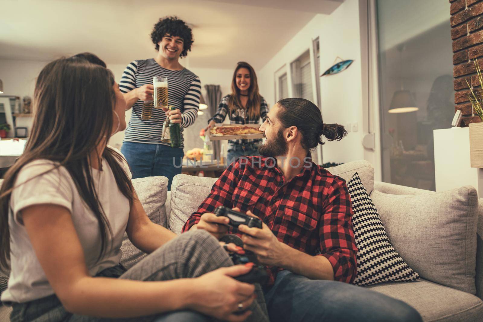 Excited couple is playing video games at home sitting on sofa. Their friends bring beer and pizza to eat together. They have great weekend in nice company indoors. 
