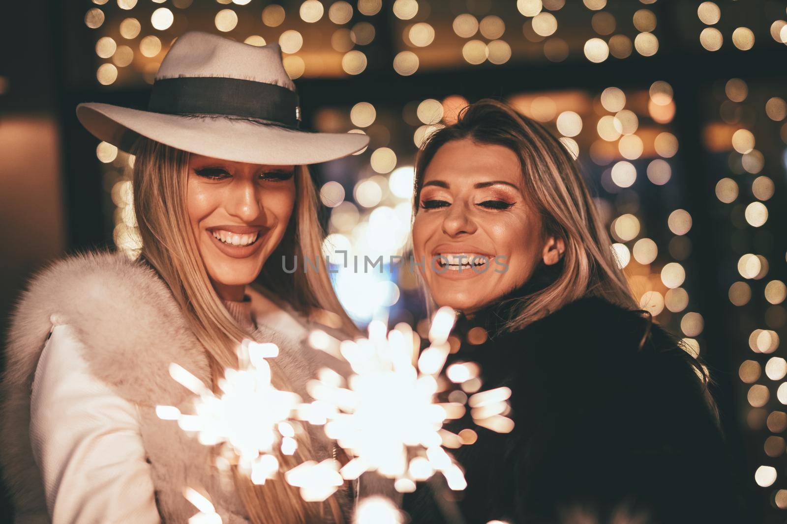 Young beautiful cheerful female friends with sparklers having fun and enjoying in the city street at Christmas eve.