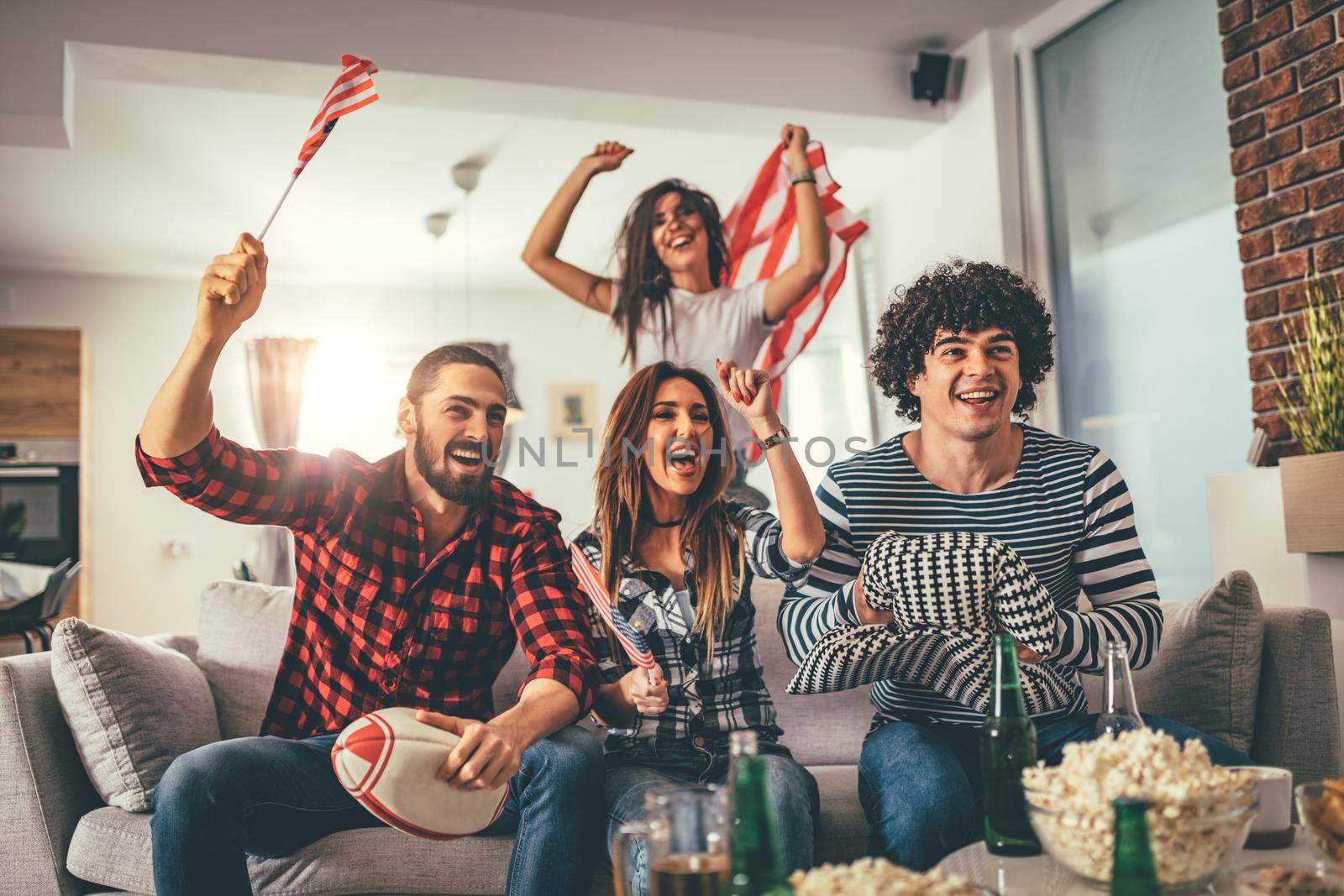 Friends are fans of sports games as American football love spending their free time at home together. They are screaming and gesturing for a victory. 