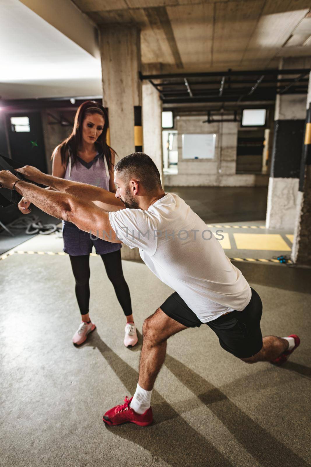 Young handsome woman having workout with a suspension straps at the gym supported of her personal male trainer. 