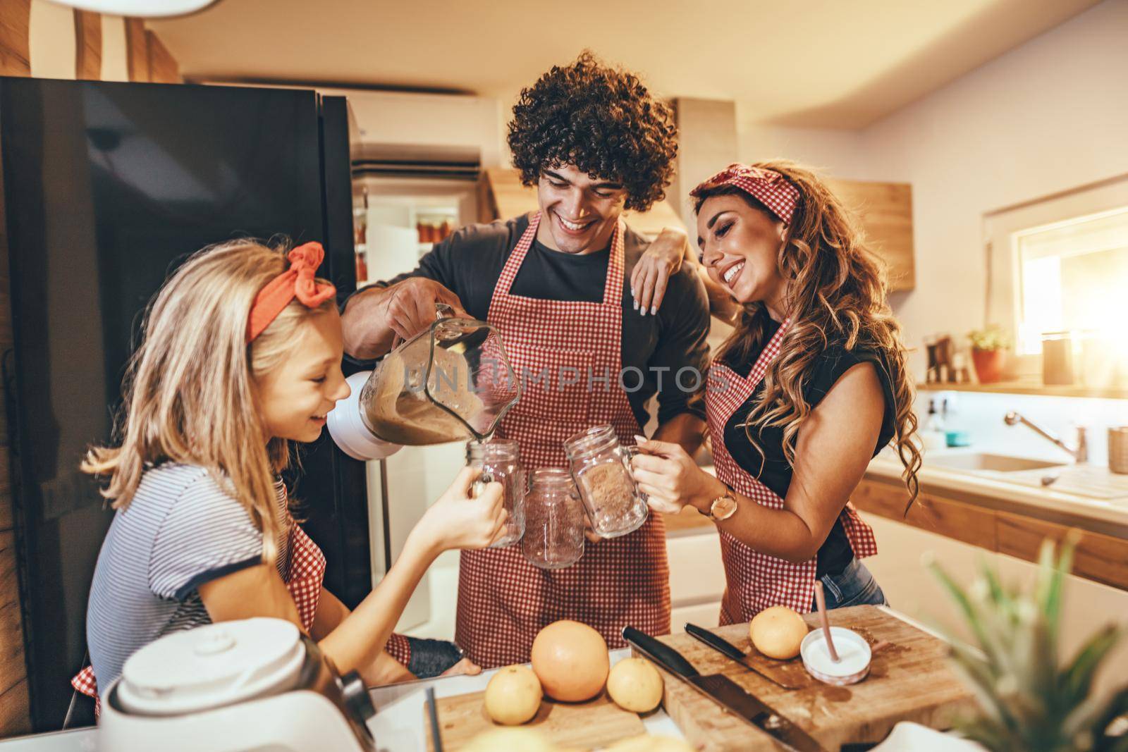 Happy young family is preparing healthy meal in the kitchen. Father is pouring cocktail into jar and mother and daughter are holding jars to drink smoothie.