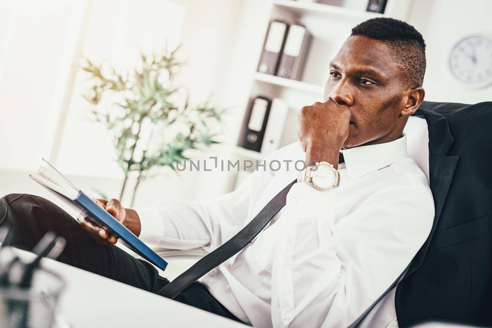 Pensive African businessman working in modern office holding notebook and planning what to do next.