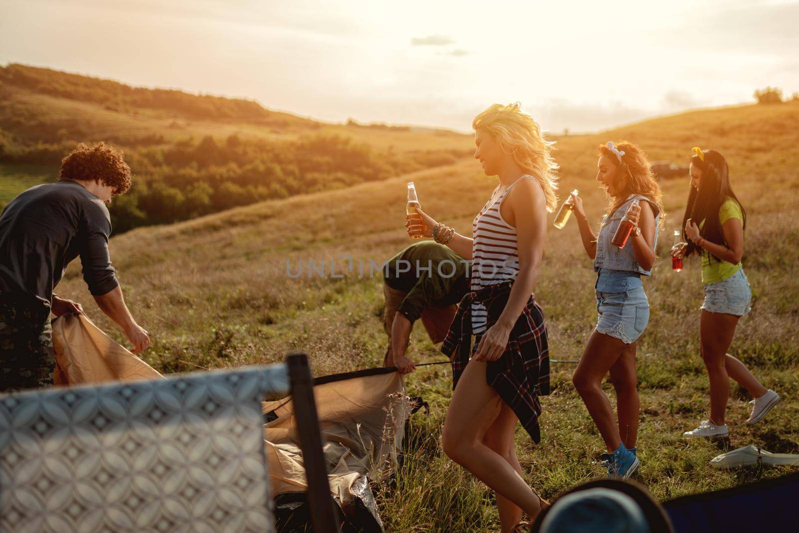 The young happy friends are preparing for camping. They're installing a tent on a suitable place in a meadow and their girlfriends are offering the beer to them.