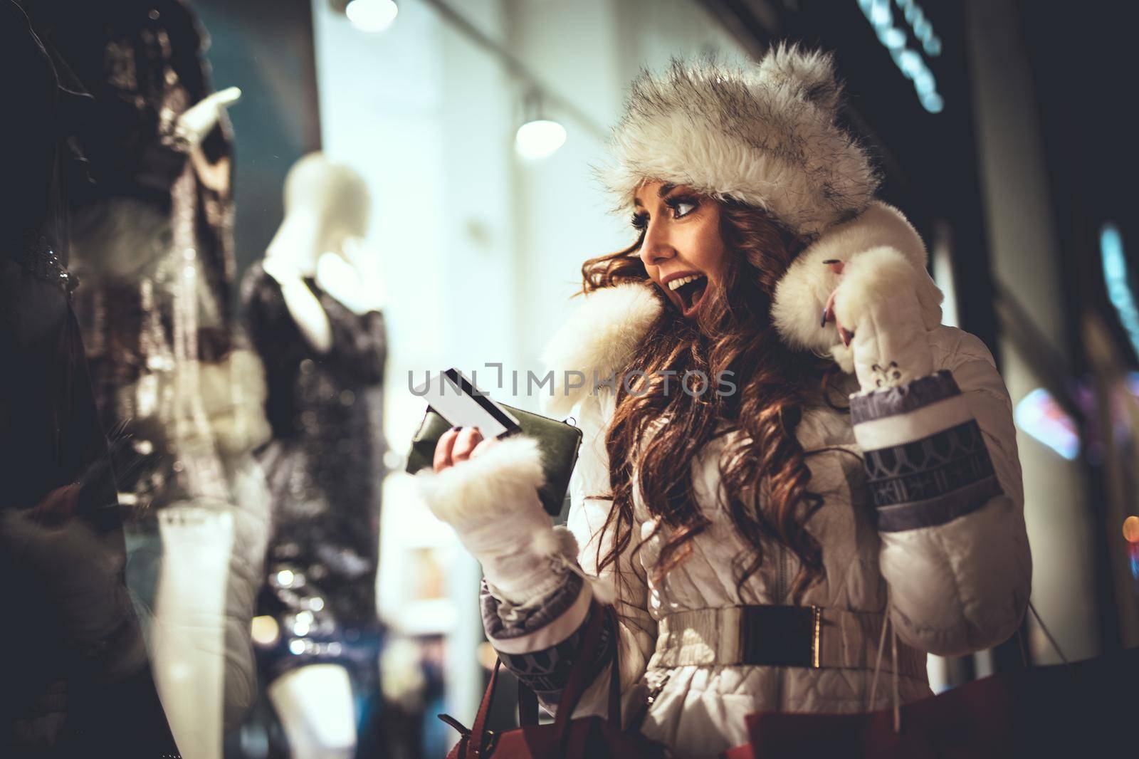 Young beautiful smiling woman having fun in the city street at Christmas time and take a photo with smartphone through the window shop.
