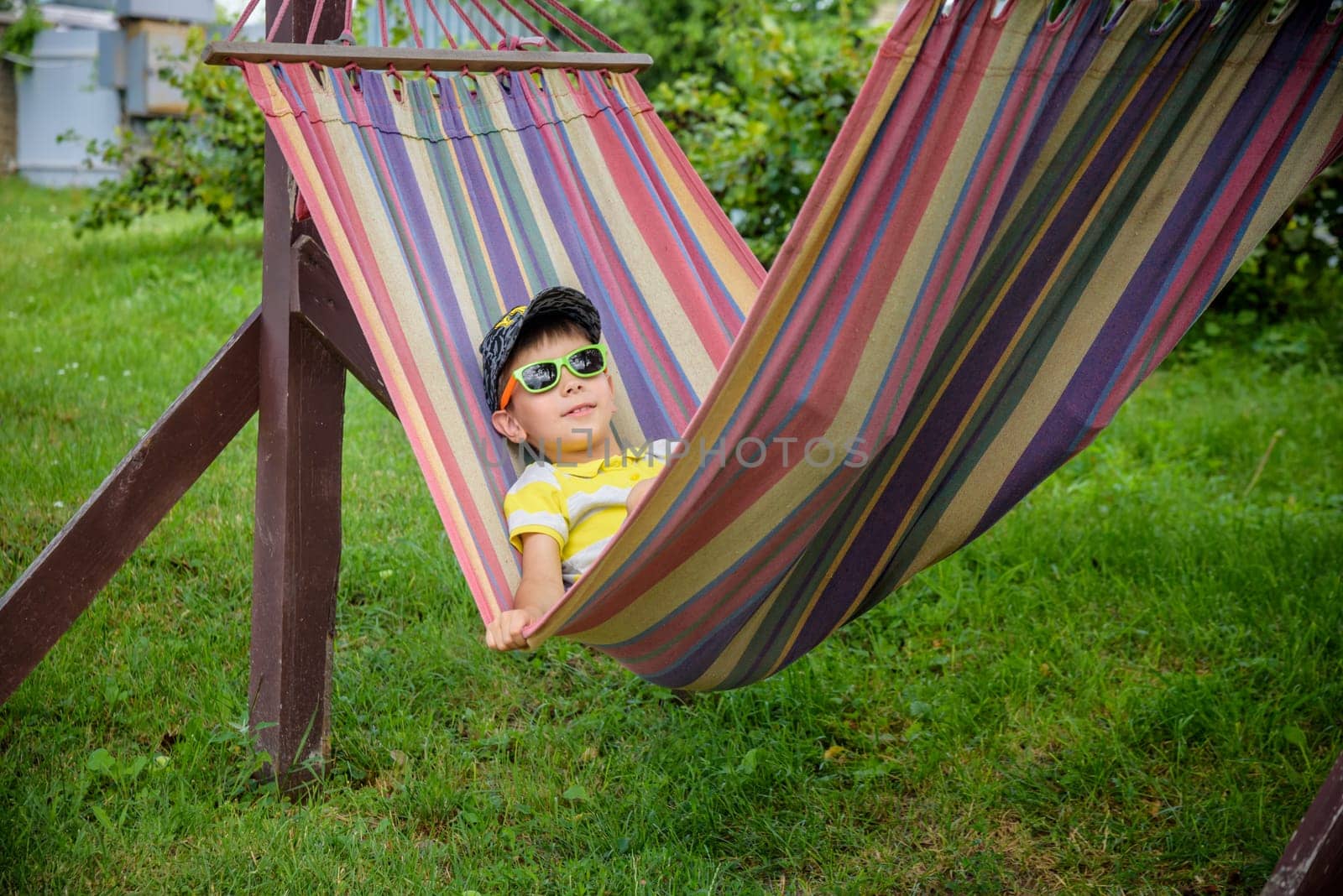 Cute little Caucasian boy relaxing and having fun in multicolored hammock in backyard or outdoor playground. Summer active leisure for kids. Child swinging on hammock. Activities for children.