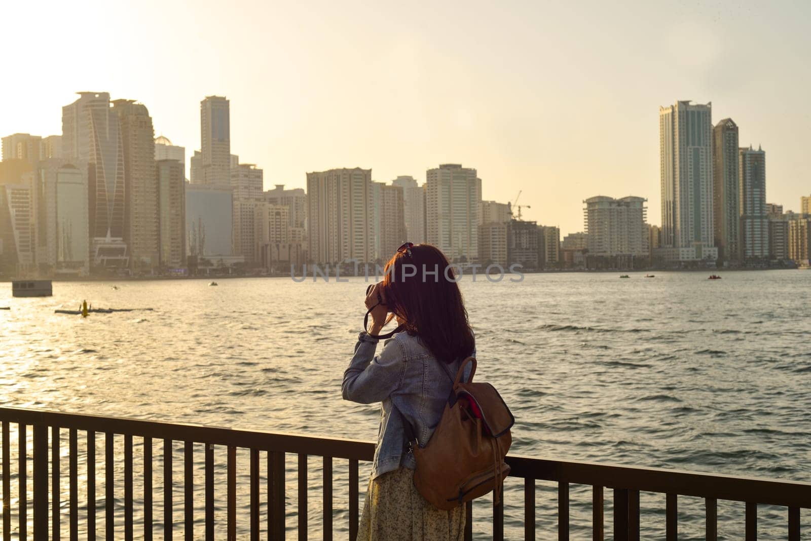 A woman in a long dress with a backpack photographs the sights on the Al Kasbah Canal in the emirate of Sharjah at sunset. Back view. by Ekaterina34