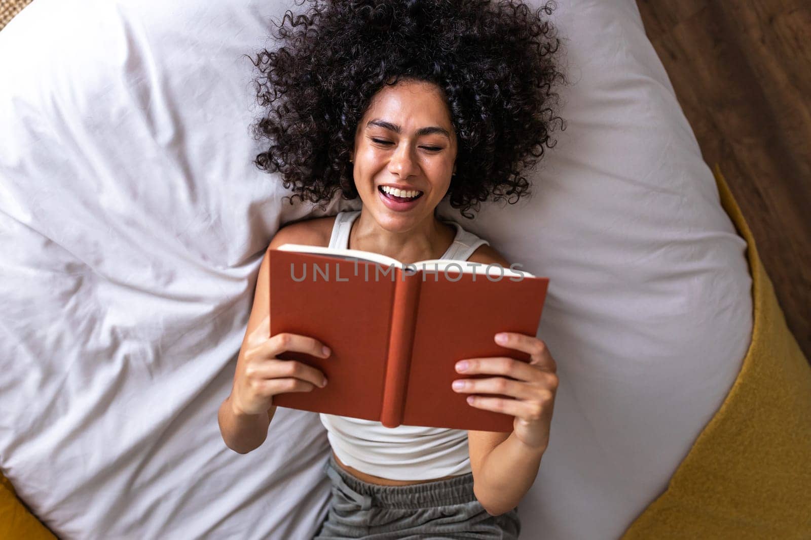 Top view of young happy multiracial woman lying on the bed reading a book laughing. by Hoverstock