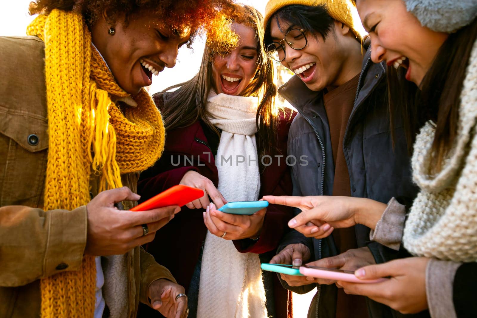 Cheerful multiracial friends looking at mobile phone together on winter day. Young woman pointing at device screen. by Hoverstock