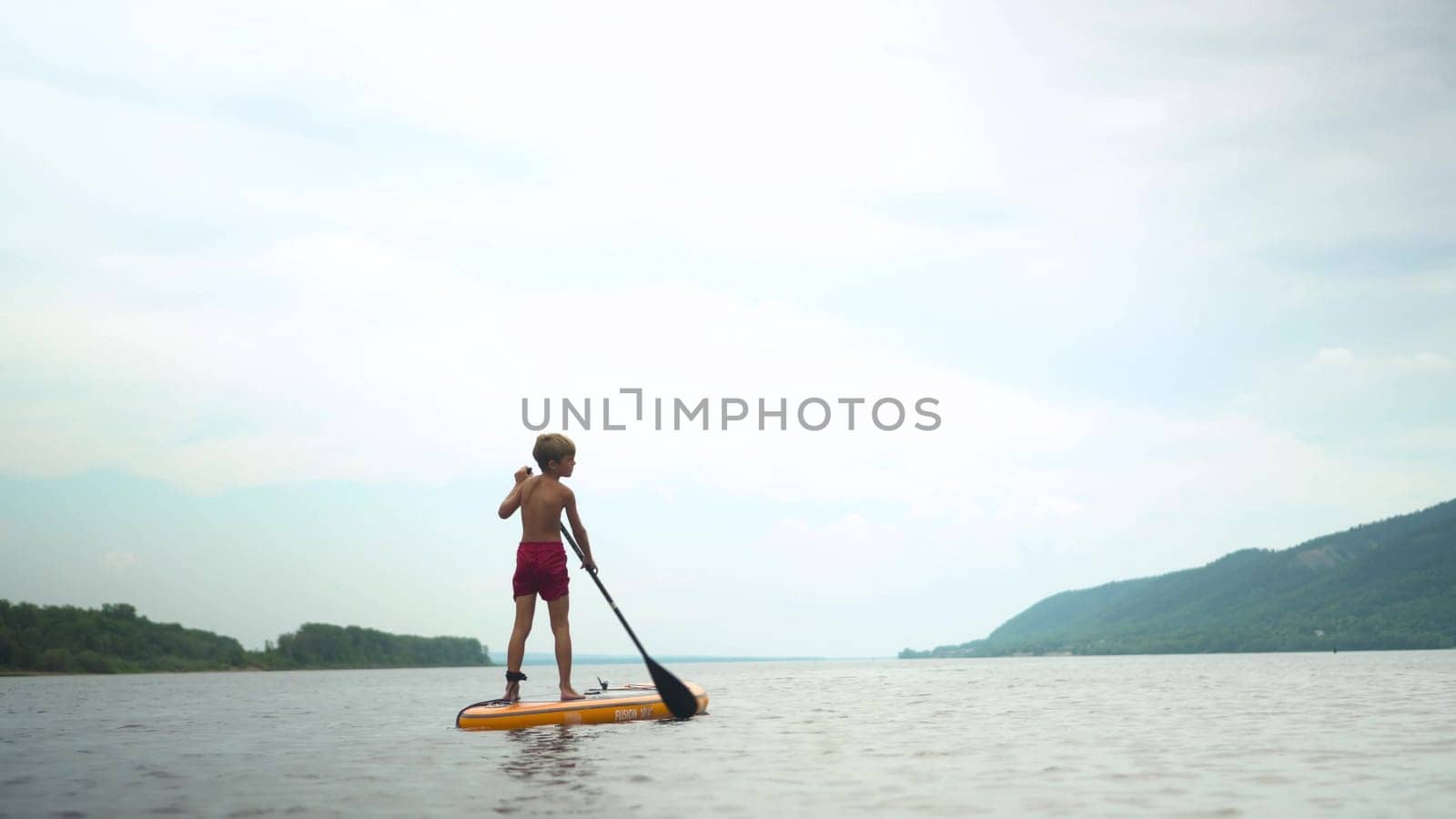 A boy on a sapsurf swims along the river against the background of the river. A sports child swims while standing on a sap. View from the back. 4k