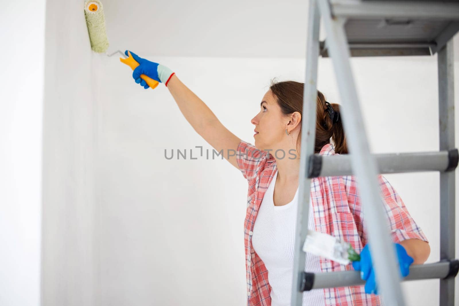 Young woman with paint roller and brush on ladder painting walls in her apartment.
