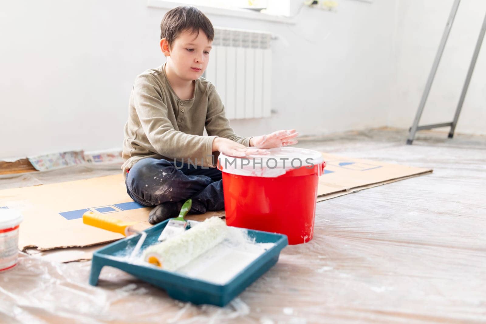 A happy child has fun sitting on the floor and banging his hands on a bucket of paint. Repair is easy. Repair in the apartment