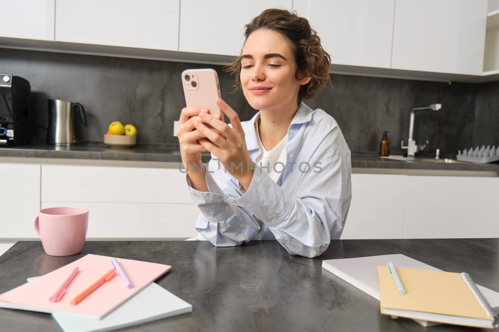 Indoor shot of beautiful woman chats on mobile phone, texts message on smartphone, orders takeaway, groceries delivery in app.