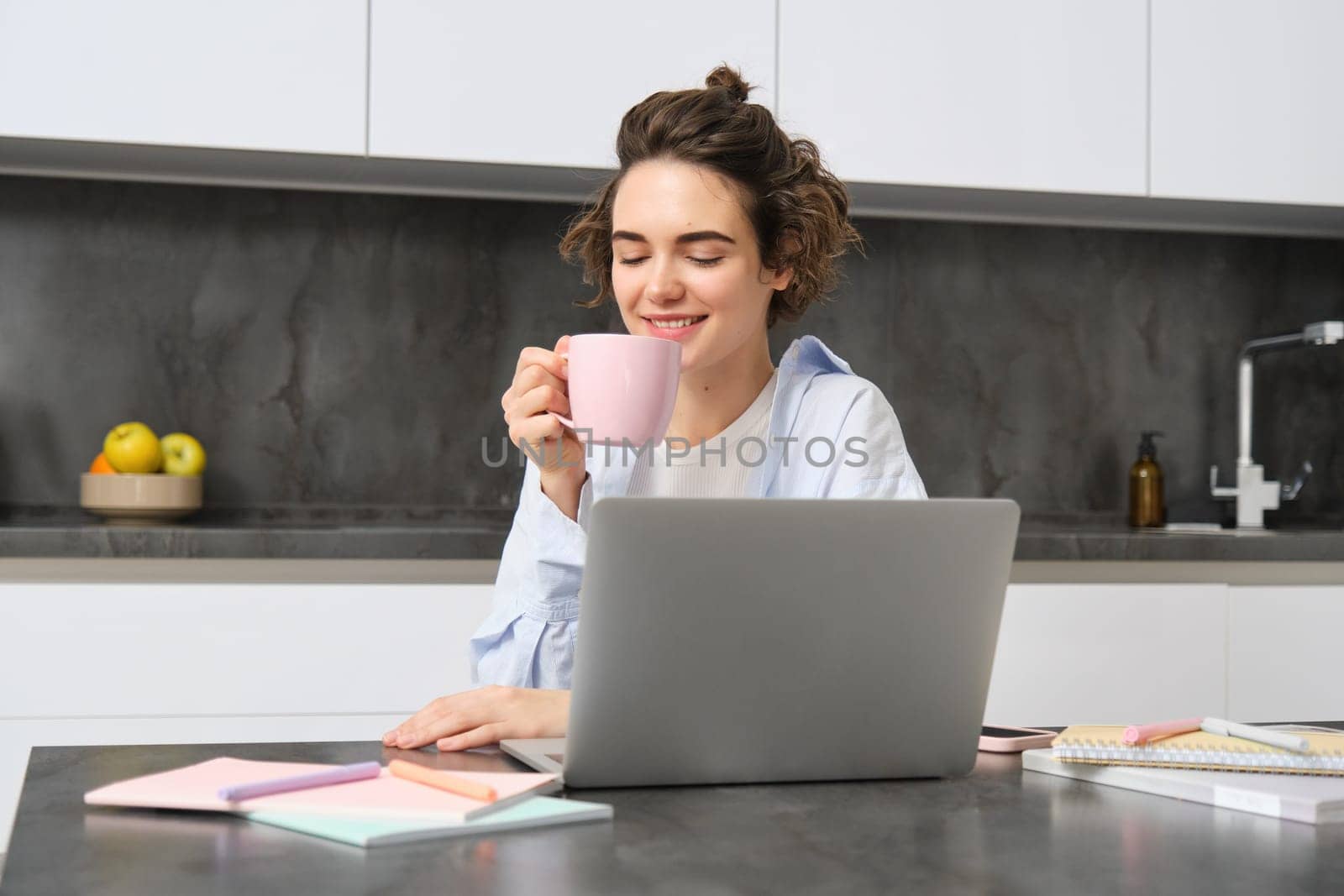 Smiling girl student, drinks coffee, studies online course from her laptop at home. Woman does her homework, works on remote, watches streaming show in internet by Benzoix