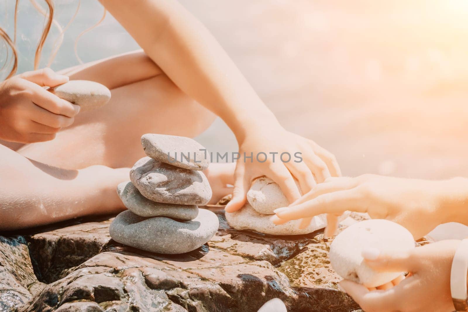 Woman with daughter bilds stones pyramid on seashore on a sunny day on the blue sea background. Happy family holidays. Pebble beach, calm sea. Concept of happy vacation on the sea, meditation, spa by panophotograph