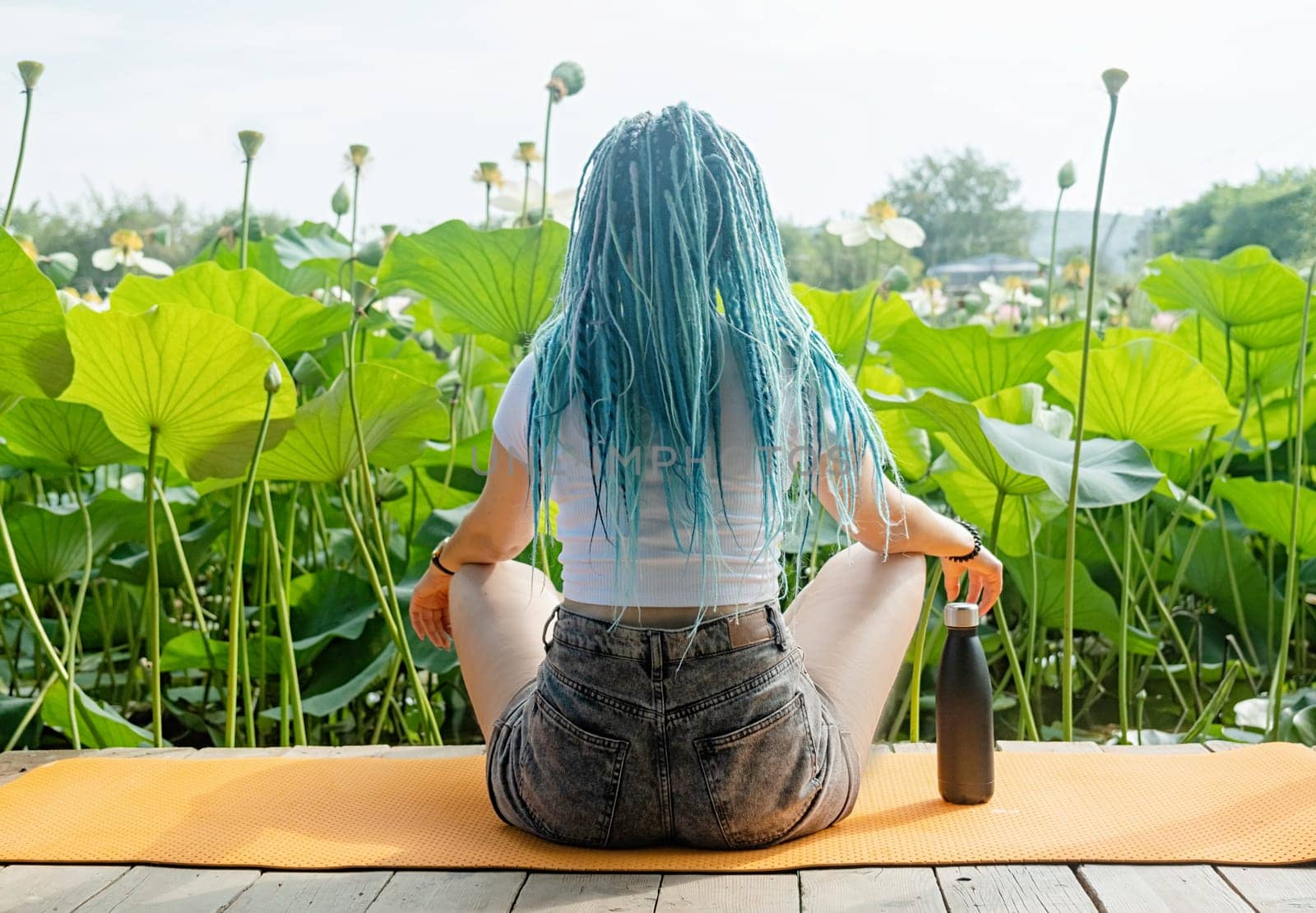 young beautiful woman with blue afro locks resting on yoga mat on wooden pierce on lotus lake enjoying nature, view from behind