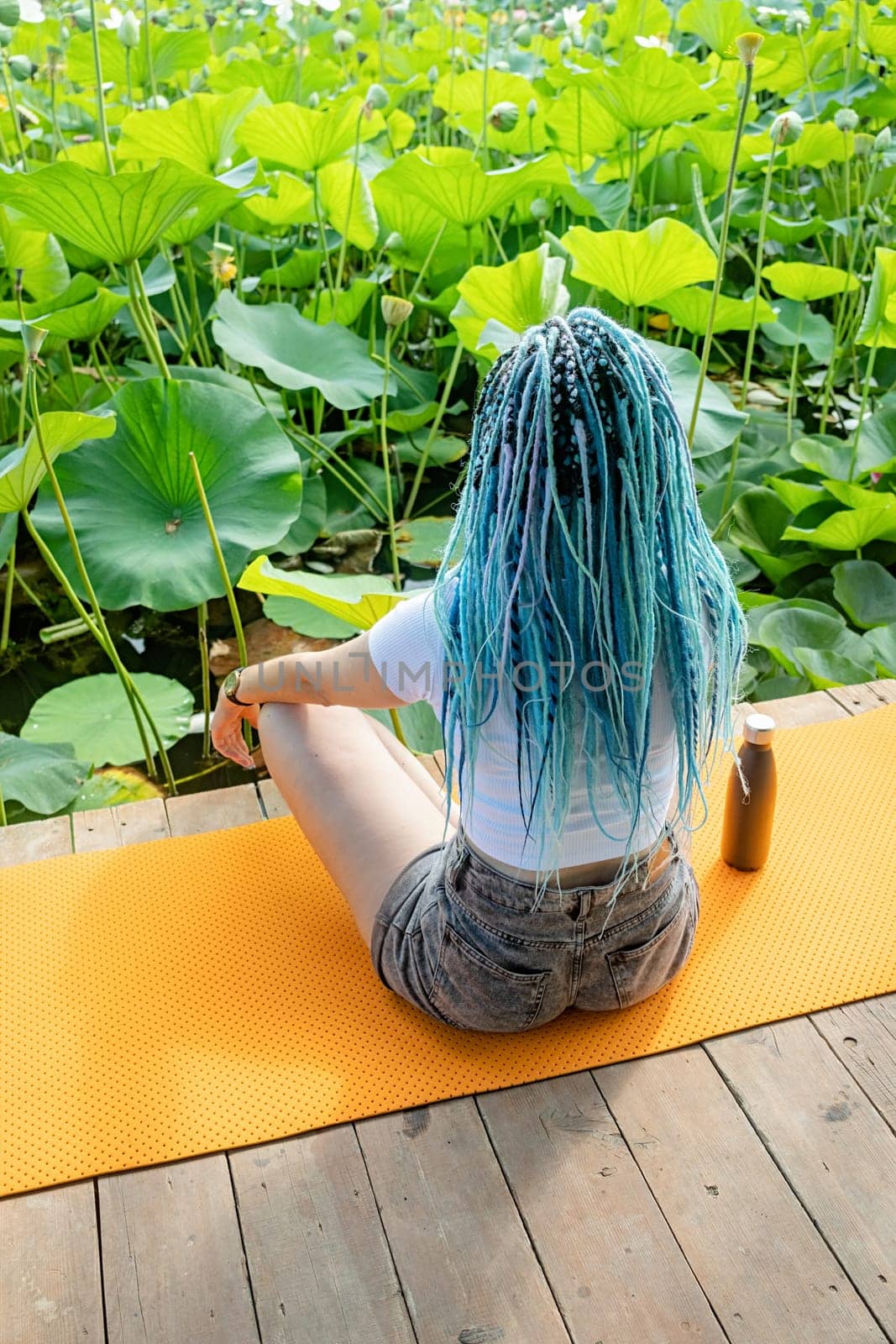 young beautiful woman with blue afro locks resting on yoga mat on wooden pierce on lotus lake enjoying nature, view from behind