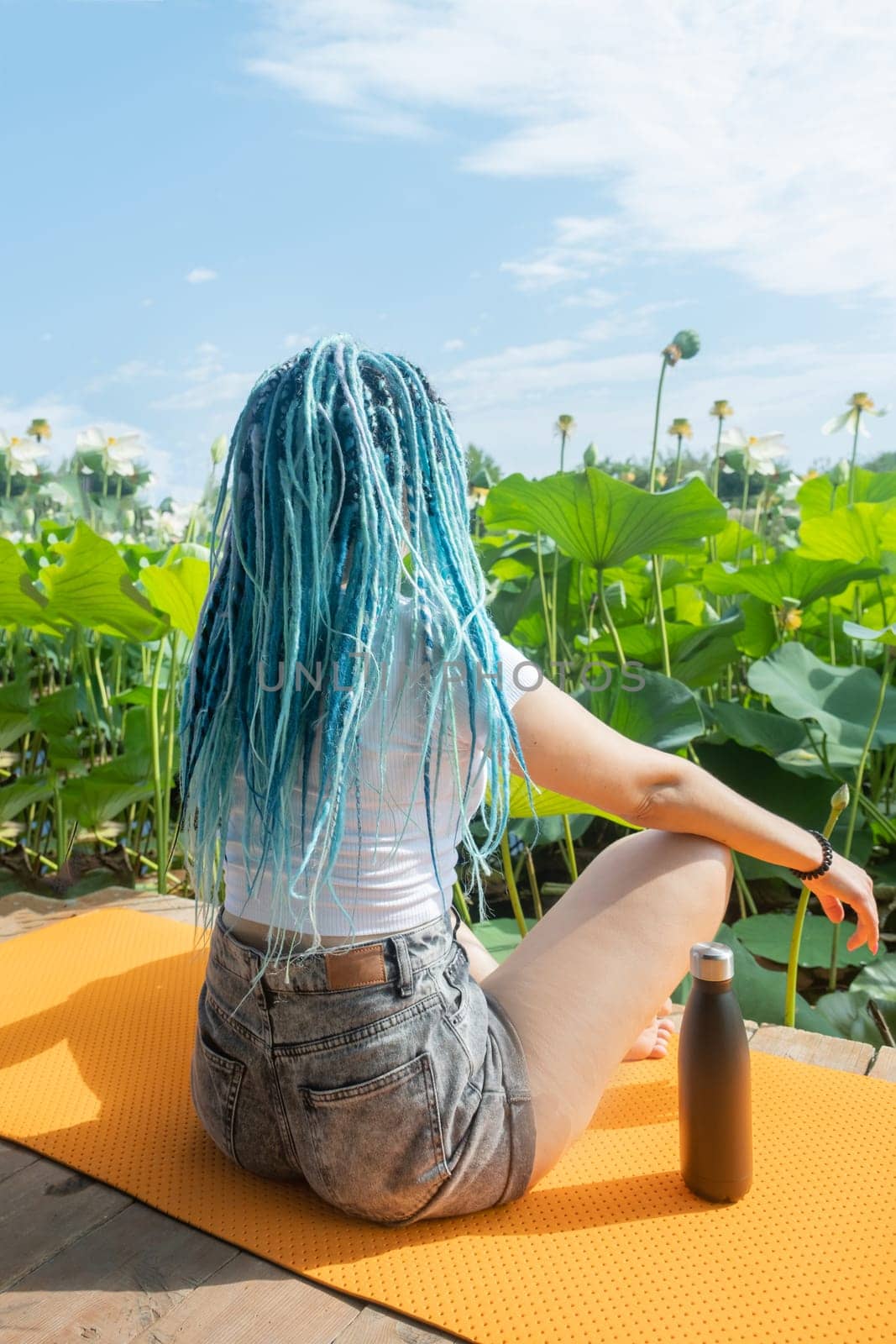 young beautiful woman with blue afro locks resting on yoga mat on wooden pierce on lotus lake enjoying nature, view from behind